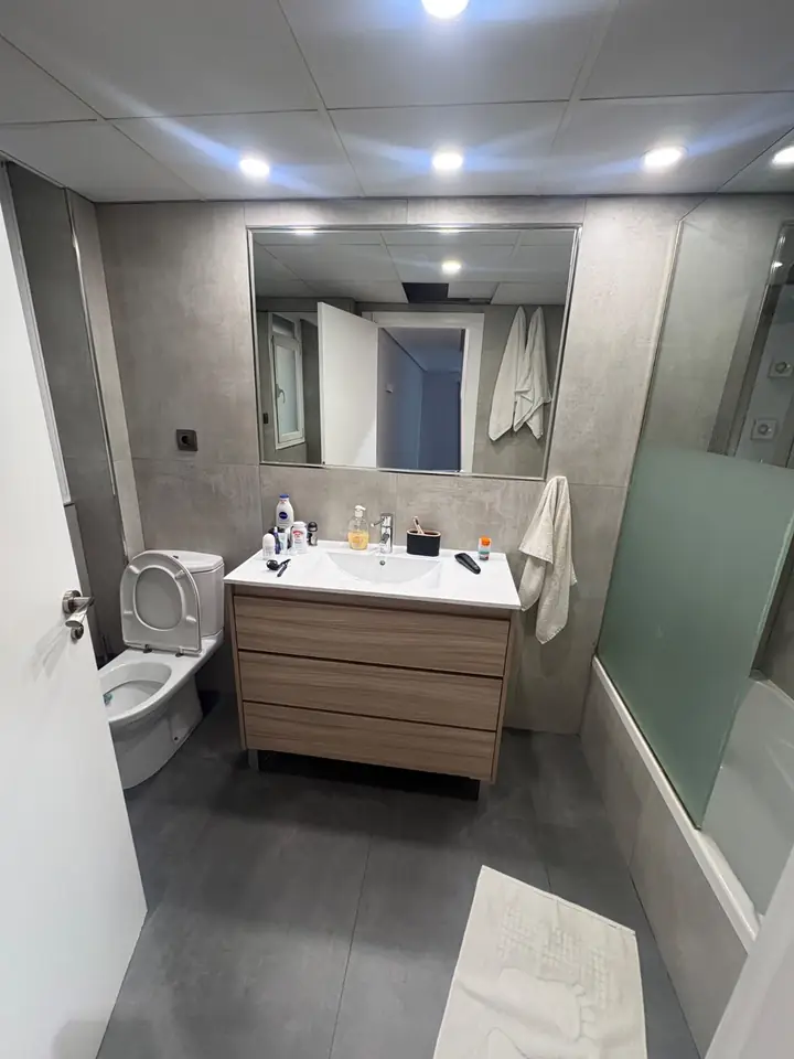 Foto real de un baño antes de la remodelación: lavatorio con mueble de madera, inodoro, bañadera con mampara y cerámica gris
